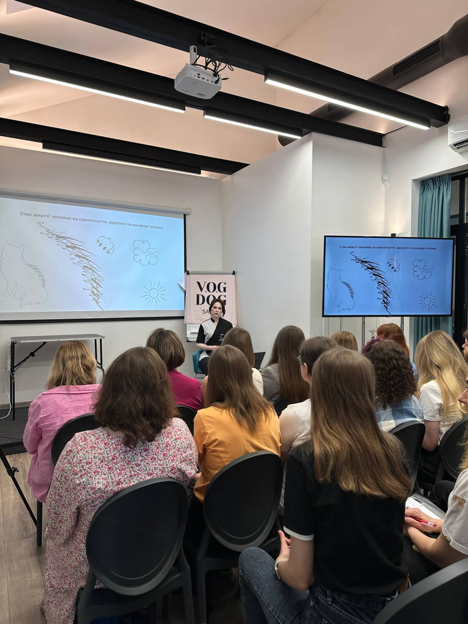 Група дівчат з різною довжиною волосся сидять перед викладачем та дивлять на екран