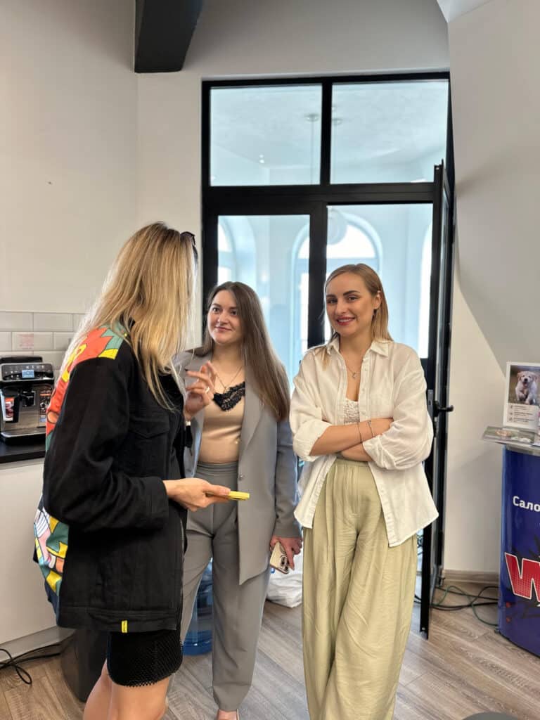 Дівчина в бежевих лляних штанах посміхається на камеру, поки інші ведуть бесіду