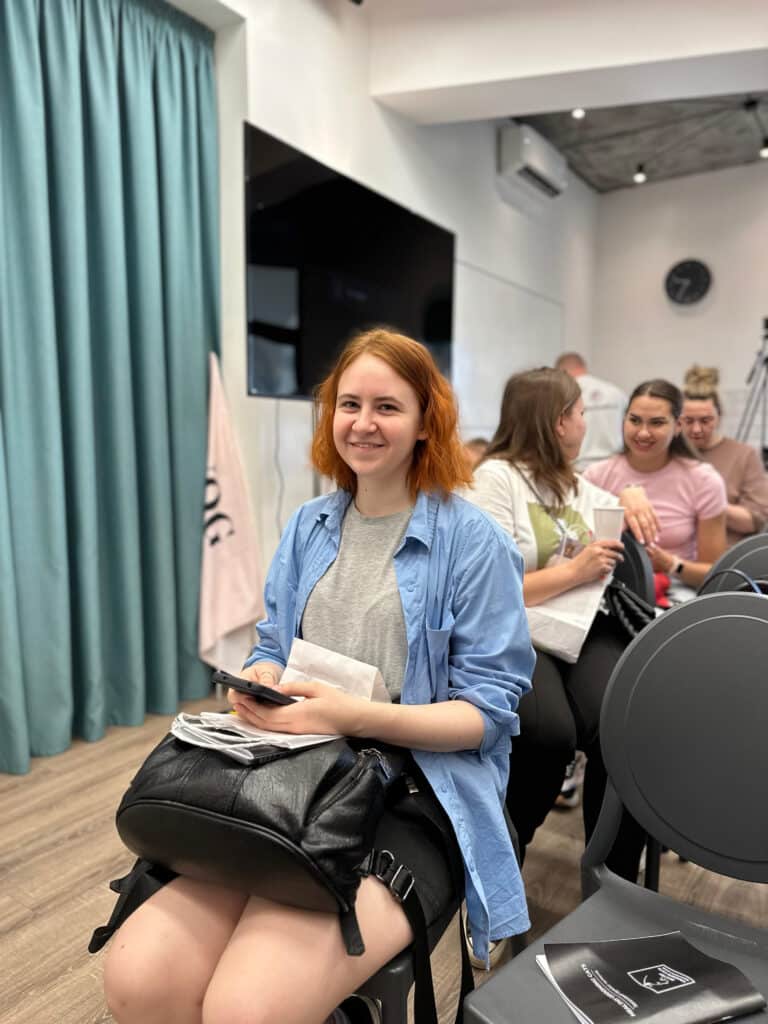 Дівчина з рудим волоссям та в синій сорочці тримає в руках телефон, а на колінах - сумку