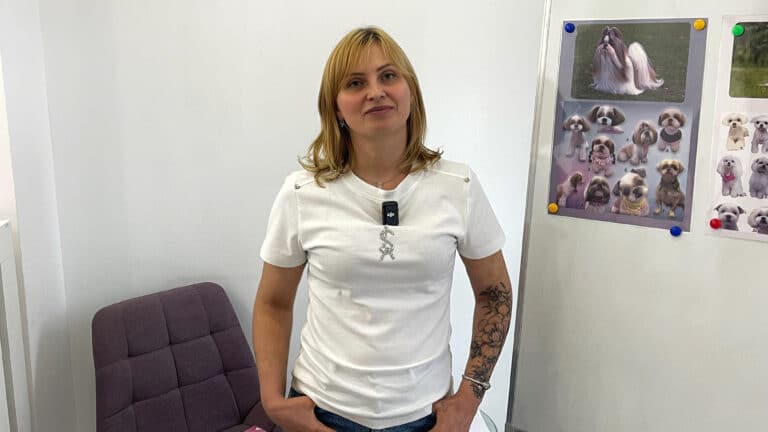 Дівчина в білій футболці стоїть біля сірого стільця в білій кімнаті