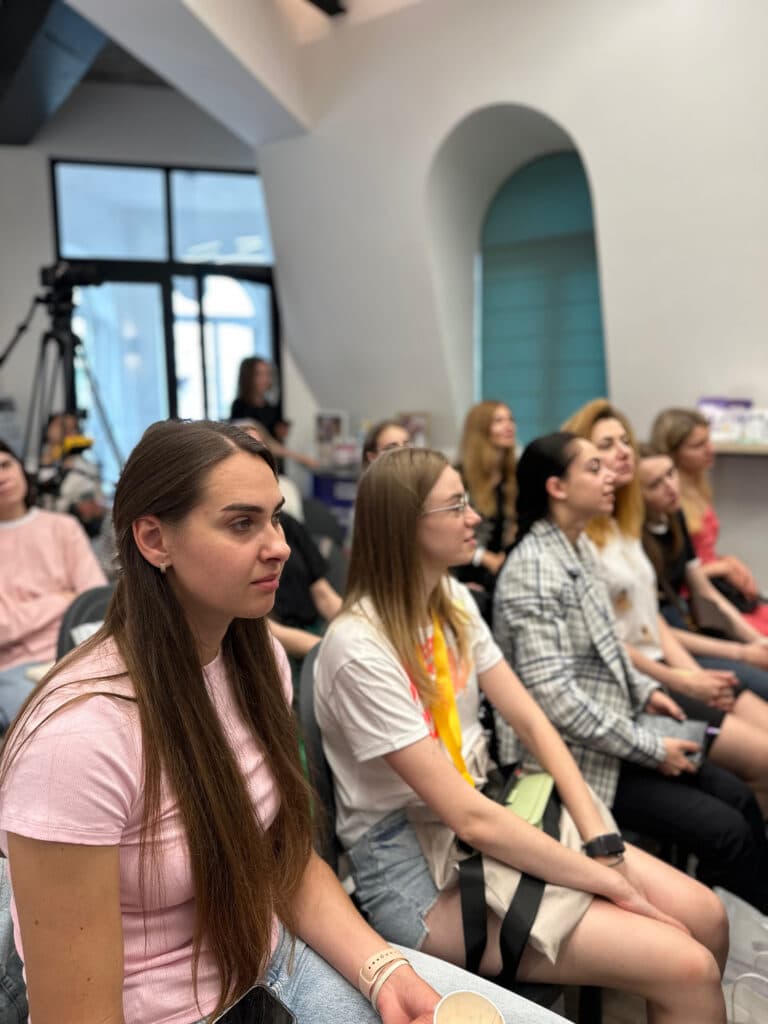 Дівчина з довгим волоссям та в рожевій футболці сидить першою і уважно слухає інформацію