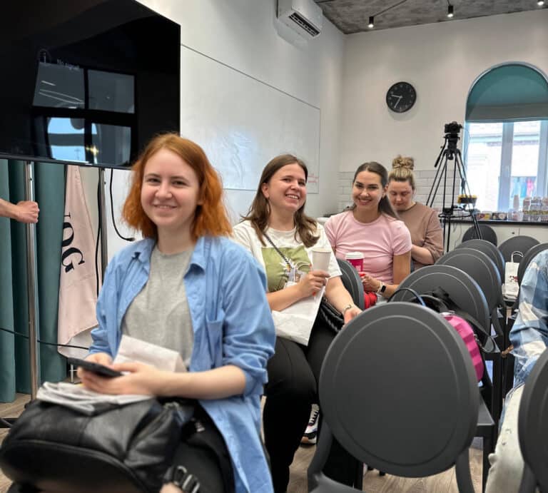 Чотири дівчини в ряд посміхаються, сидячи в одному приміщенні для проведення курсів