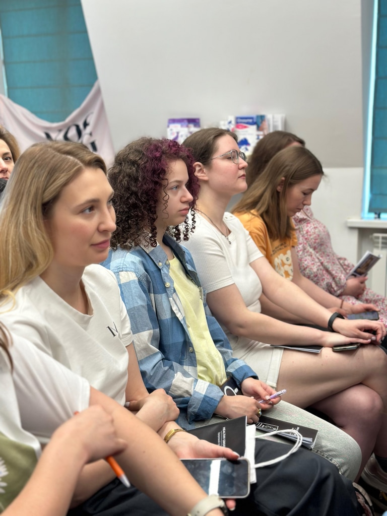 Дівчата сидять на стільцях в першому ряду, тримаючи записники та ручки