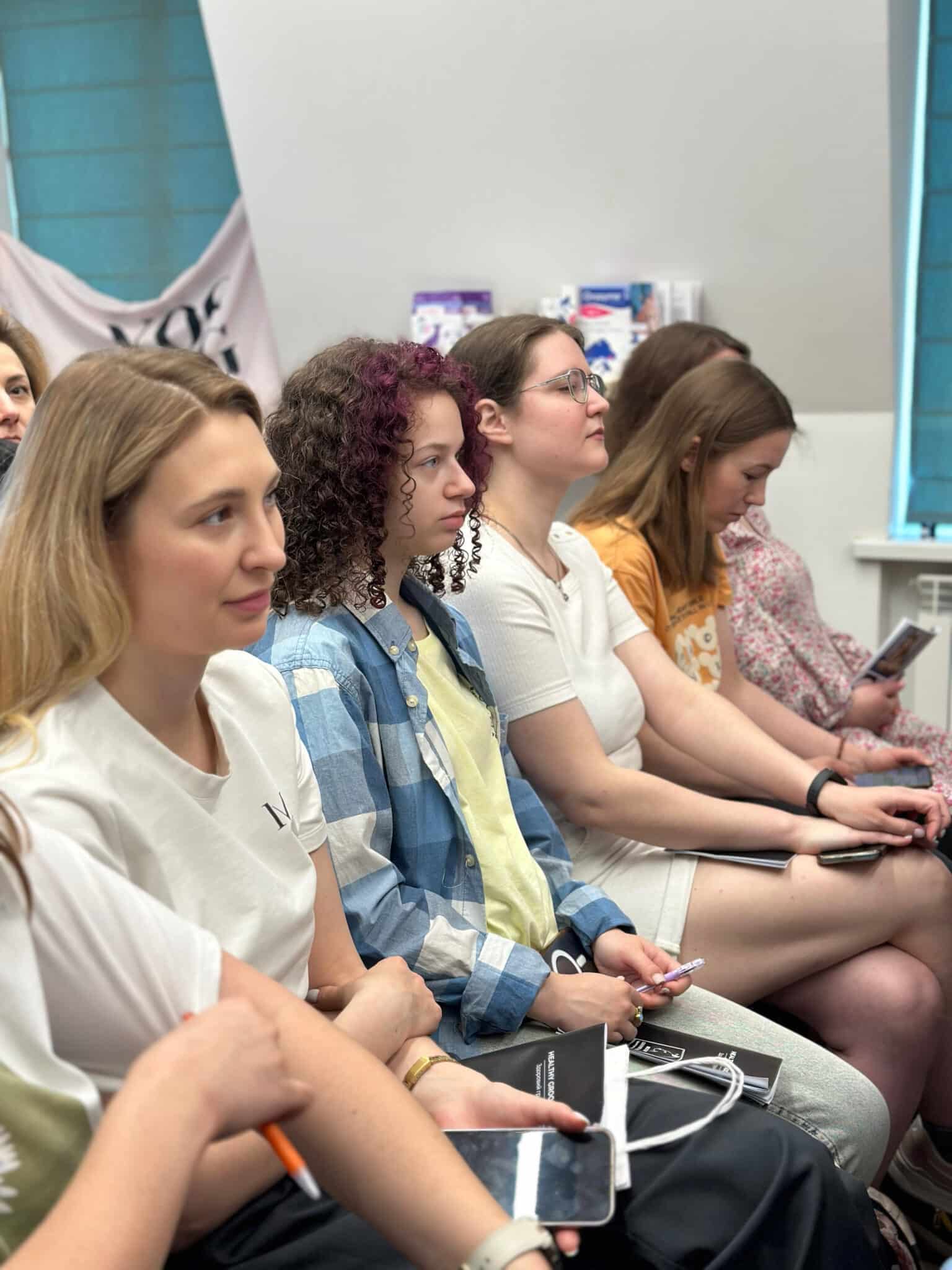 Дівчата сидять на стільцях в першому ряду, тримаючи записники та ручки