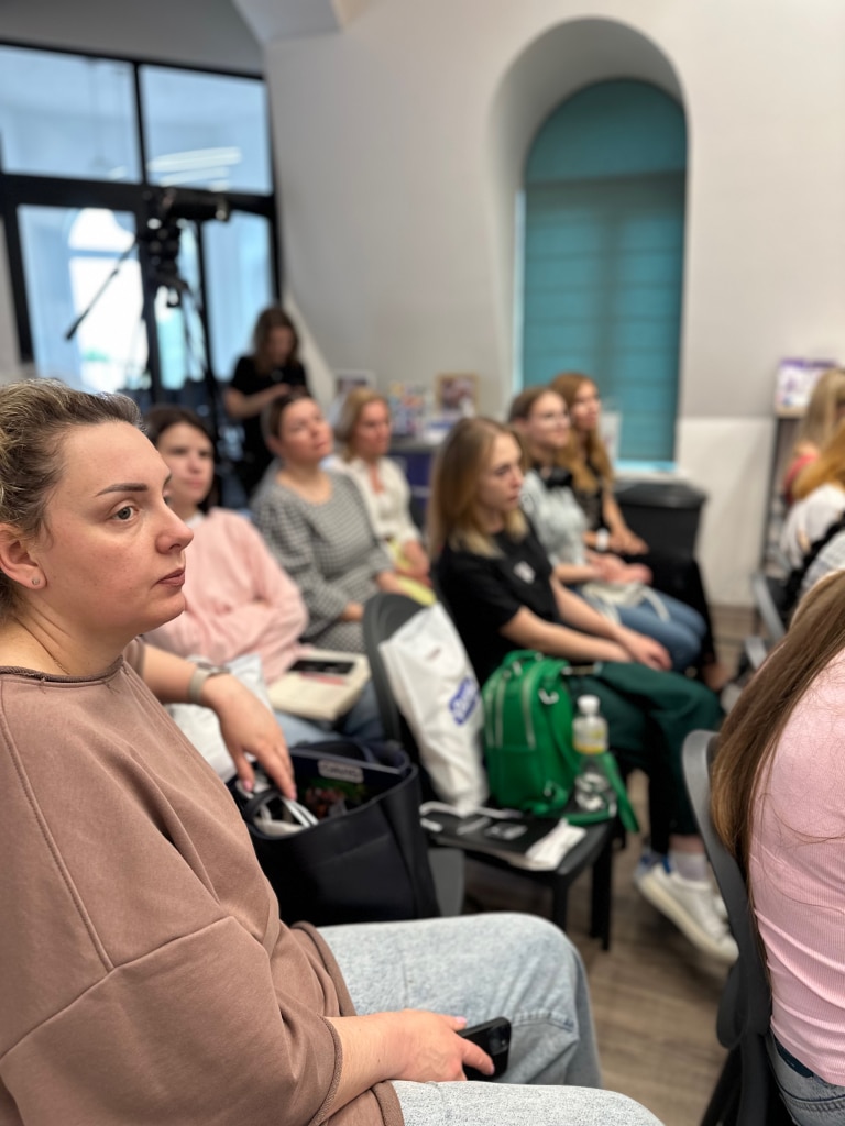 Дівчина в бежевій довгій футболці сидить біля двох пустих стільців