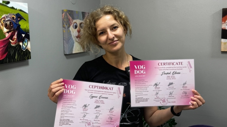 Жінка в чорній футболці тримає в кожній руці по сертифікату з грумінгу