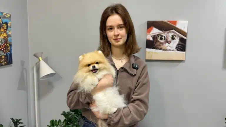 Дівчина з маленьким шпіцом на руках стоїть поруч з білим мінімалістичним світильником