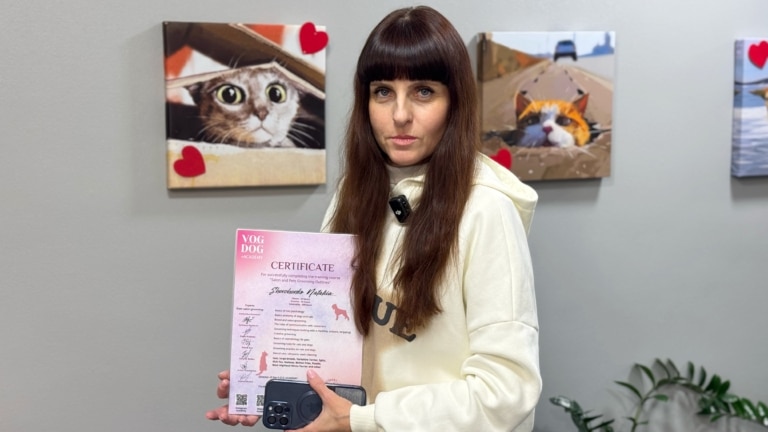 Дівчина з довгим волоссям та вкладеним чубчиком тримає в руках сертифікати рожевого кольору і телефон