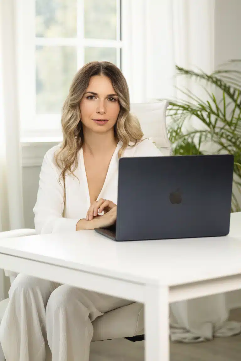 Жінка у білій блузі та штанах сидить за білим столом на білому кріслі