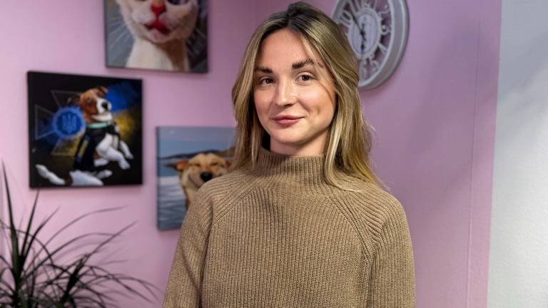 Дівчина у бежевому светрі стоїть спиною до картин та білого годинника