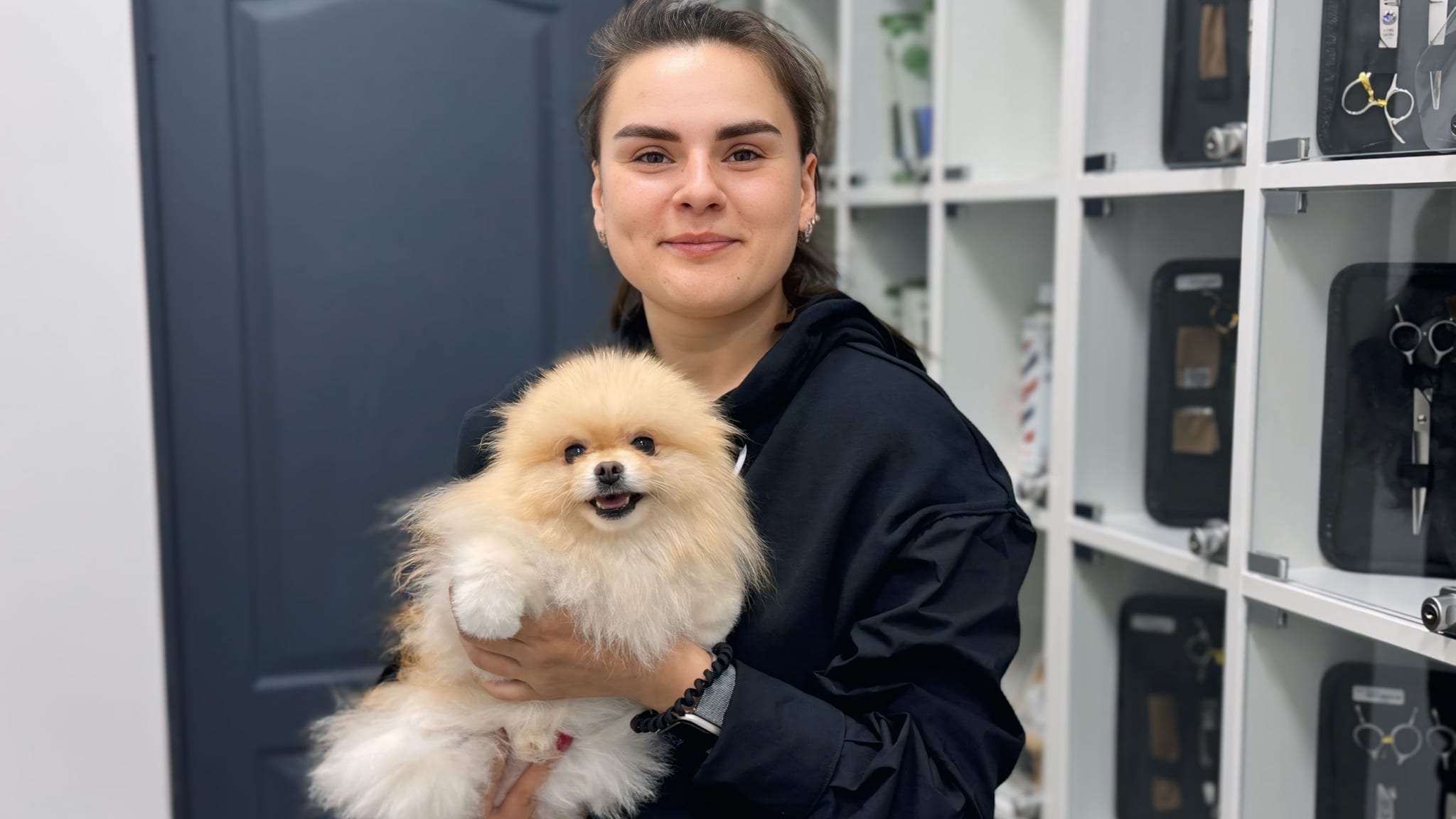 Дівчина в чорному одязі тримає в руках маленького кремового шпіца біля полиць з інструментом