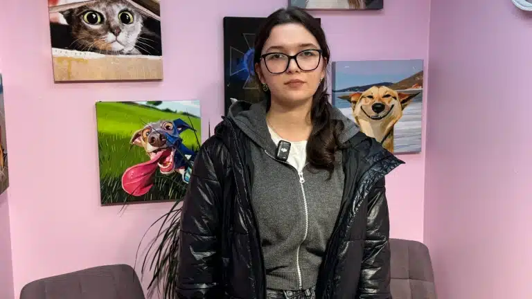 Дівчина в чорних окулярах та лаковій куртці має позаду себе картини з тваринами